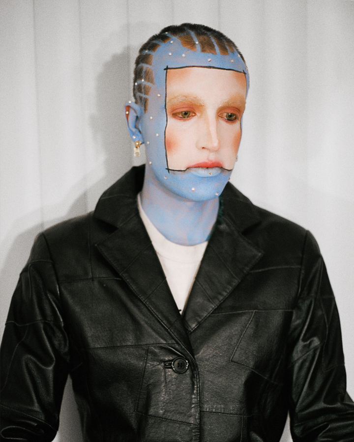 Waist-up photo of a model with blue skin and and white square in the center of their face in a black leather jacket on a white background